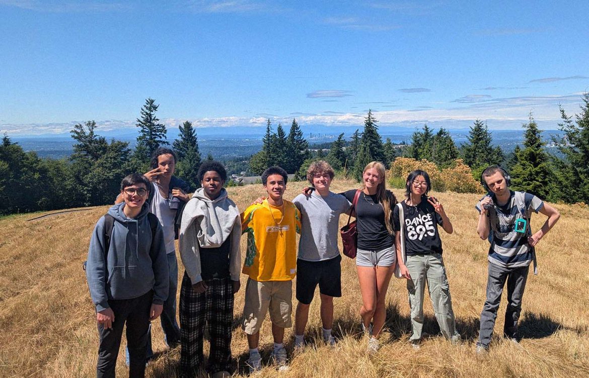 A group of people stands on a grassy hilltop with evergreen trees and a distant city skyline visible in the background under a clear blue sky.