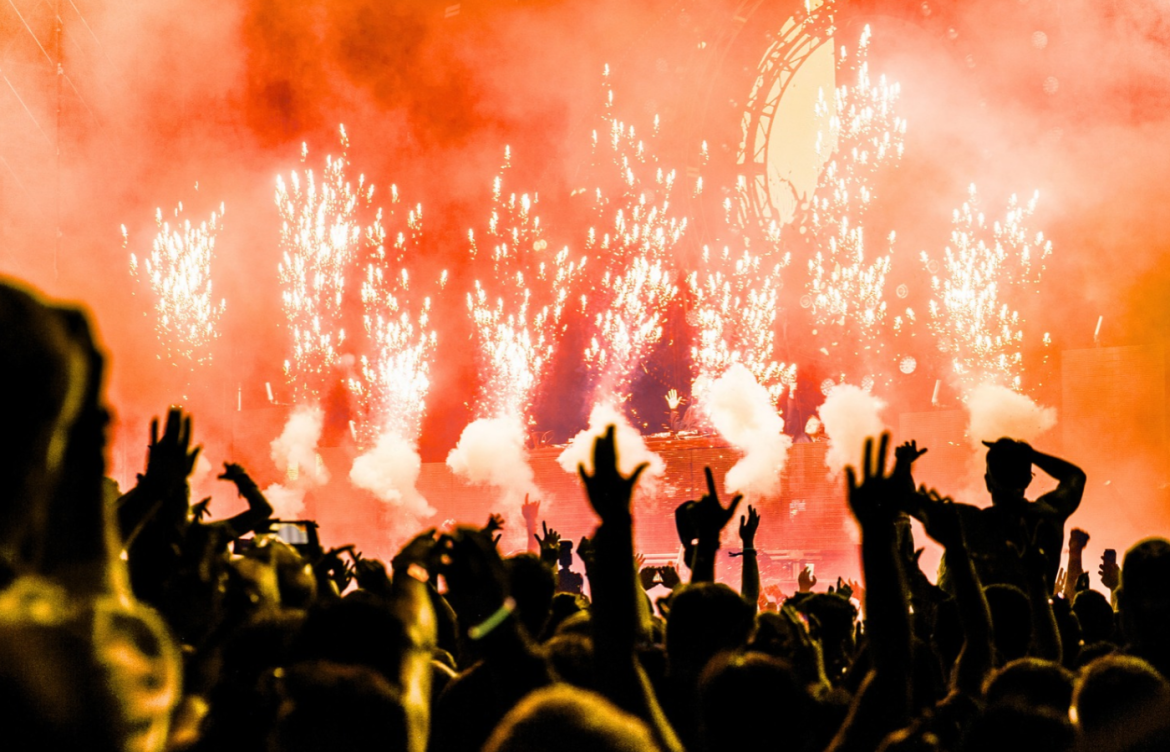 The image shows a large crowd of people at an outdoor concert, silhouetted by bright, colorful fireworks and stage lights. A DJ or performer is on stage, surrounded by vibrant pyrotechnics and smoke effects, creating an energetic atmosphere.